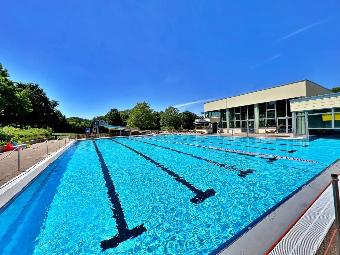 Ein Freibad mit markierten Bahnen unter strahlend blauem Himmel, neben einem modernen Gebäude, umgeben von grünen Bäumen.