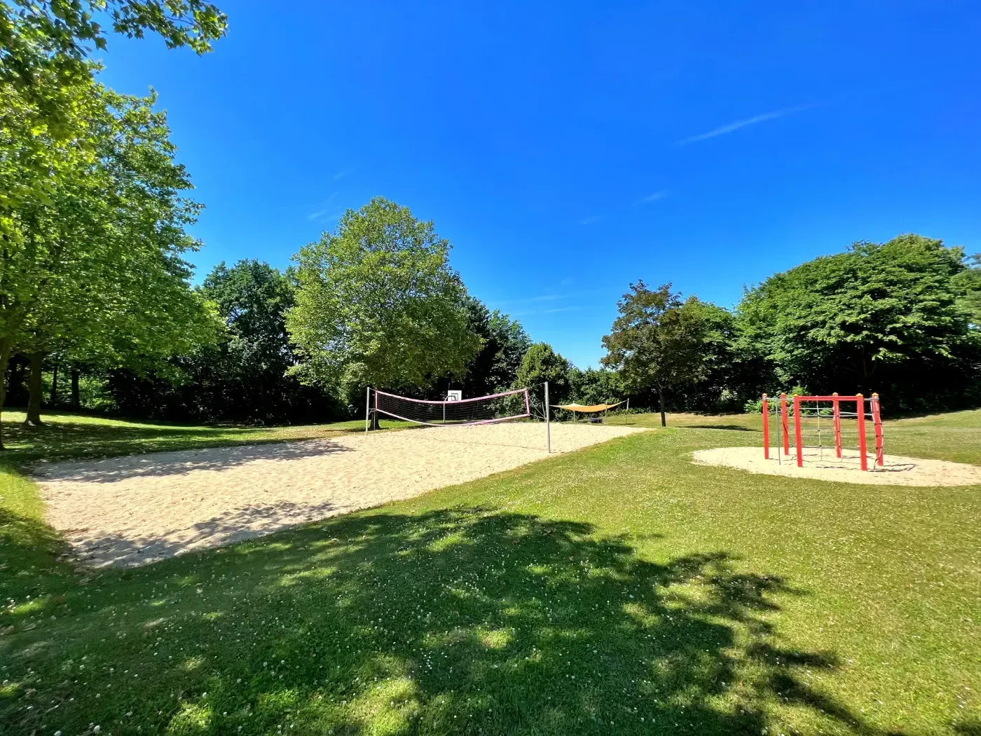 Ein sonniger Park mit einem Sandvolleyballfeld auf der linken Seite und einem roten Klettergerüst auf der rechten Seite, eingebettet in eine Rasenfläche.