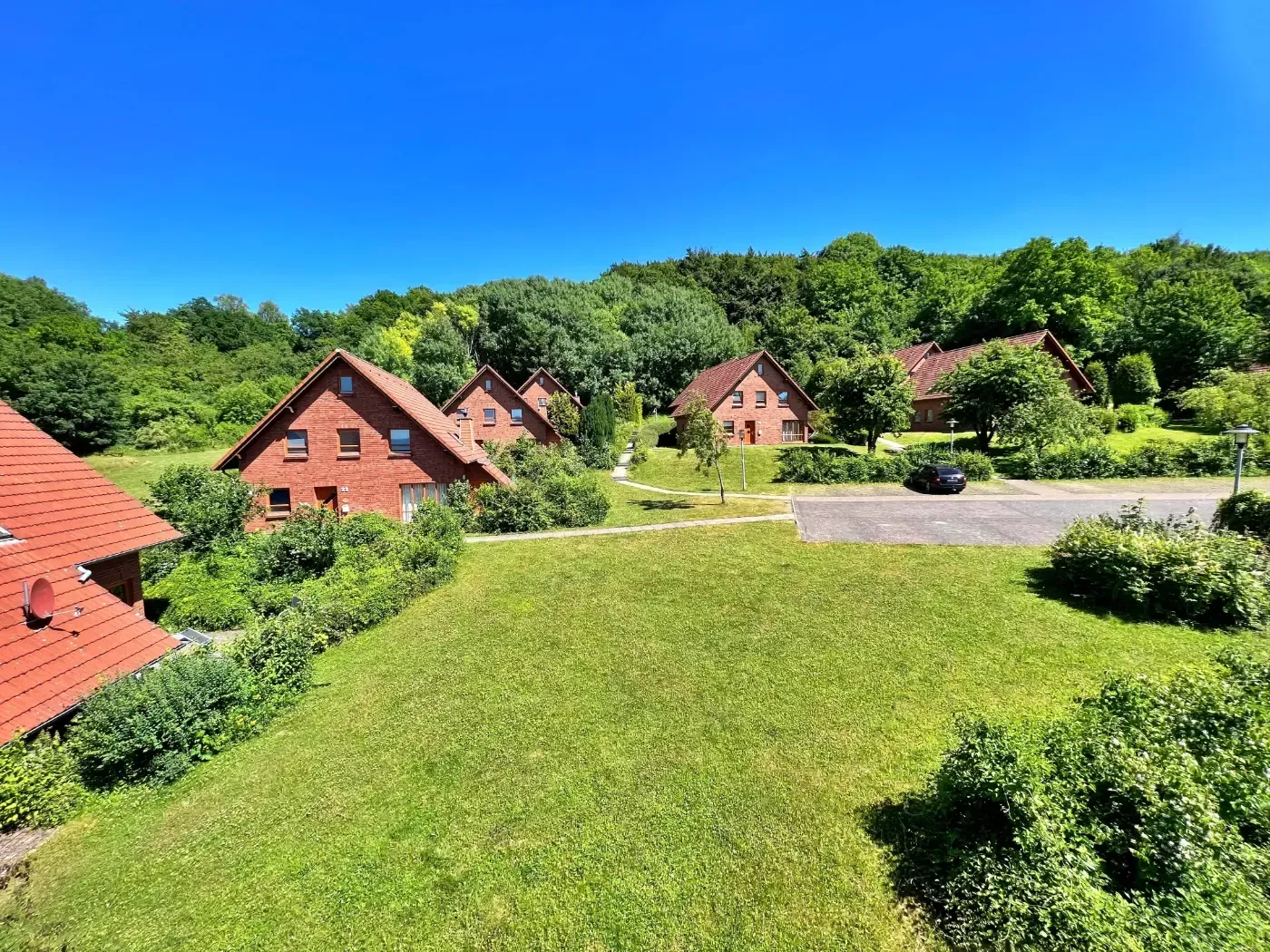 Ein strahlend sonniger Blick auf Backsteinhäuser mit roten Ziegeldächern, eingebettet in eine üppig grüne Landschaft, umgeben von dichtem Wald.