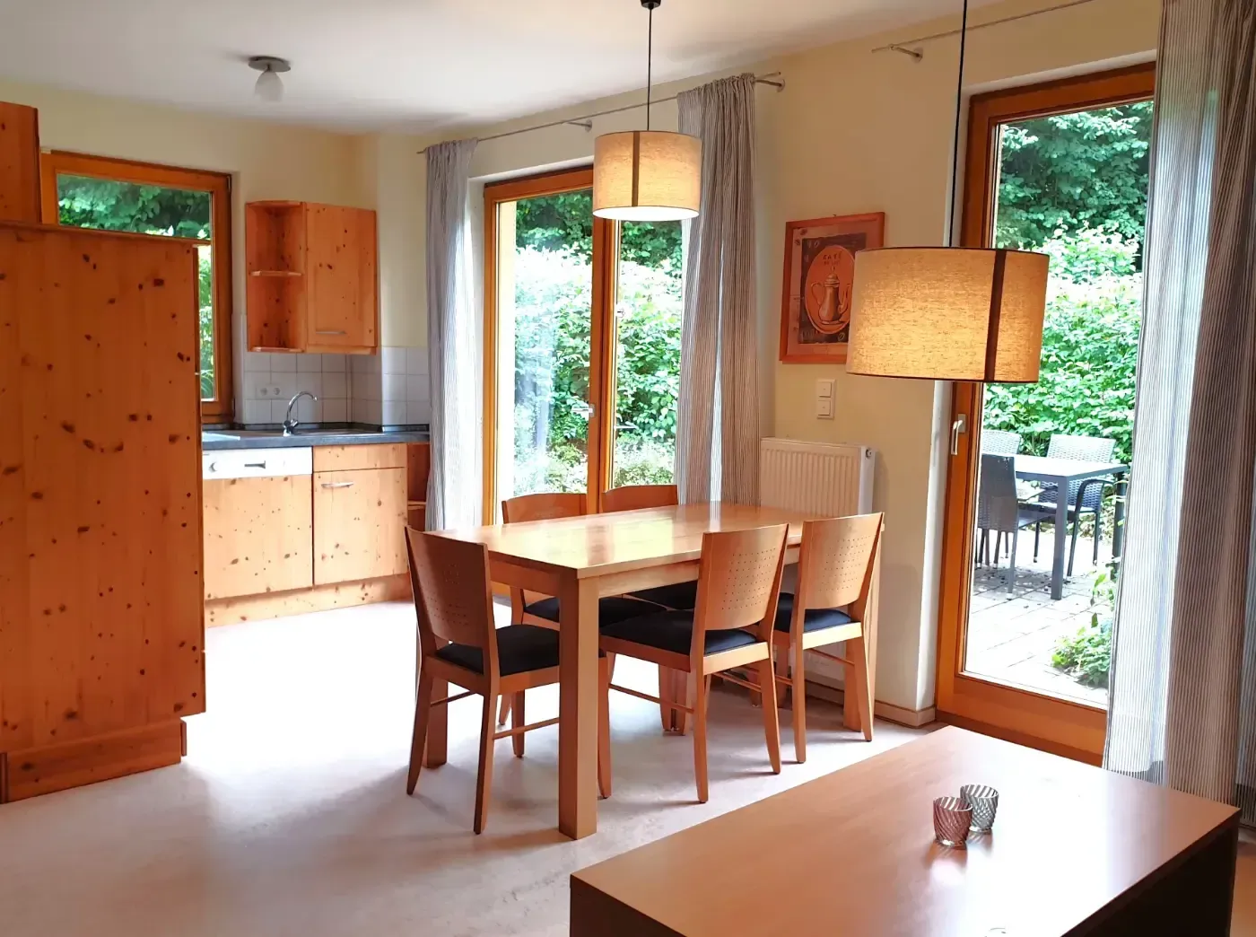 Ein Essbereich mit Holztisch, Stühlen und Lampen, neben einer Küche mit Holzschränken und einer Terrassentür.
