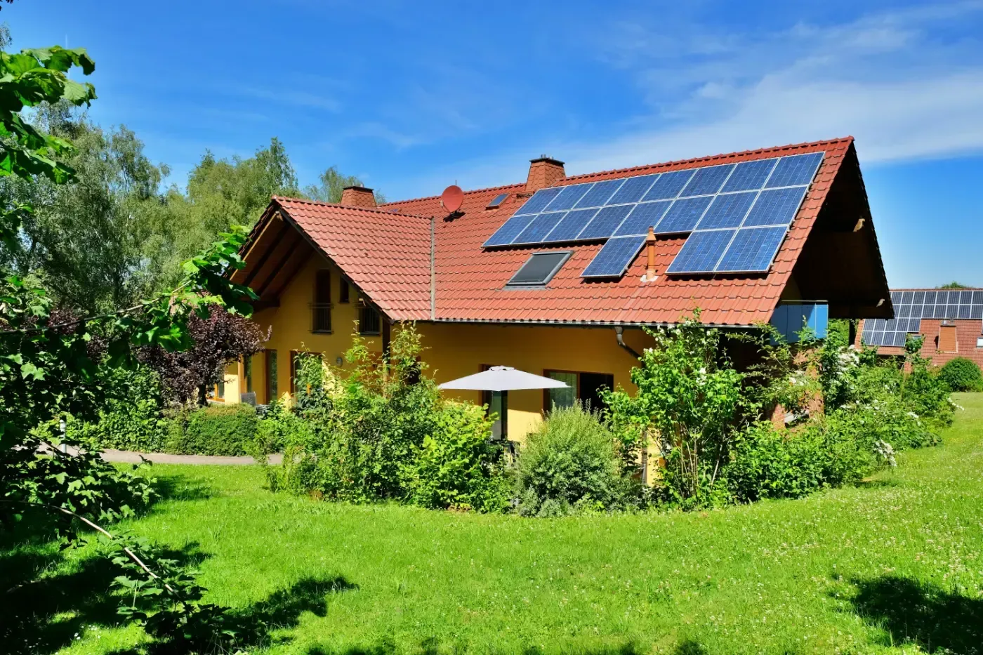 Ein gelbes Haus mit rotem Ziegeldach und Solarpaneelen, umgeben von einem üppigen grünen Garten unter strahlend blauem Himmel.