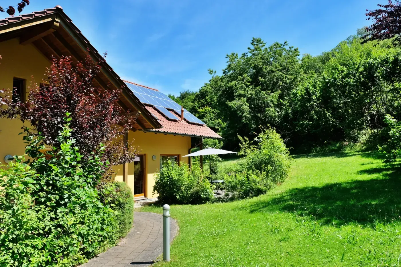 Ein gelbes Haus mit Solarpaneelen auf dem Dach, umgeben von einem üppigen grünen Rasen, Bäumen und einem gepflasterten Gehweg.