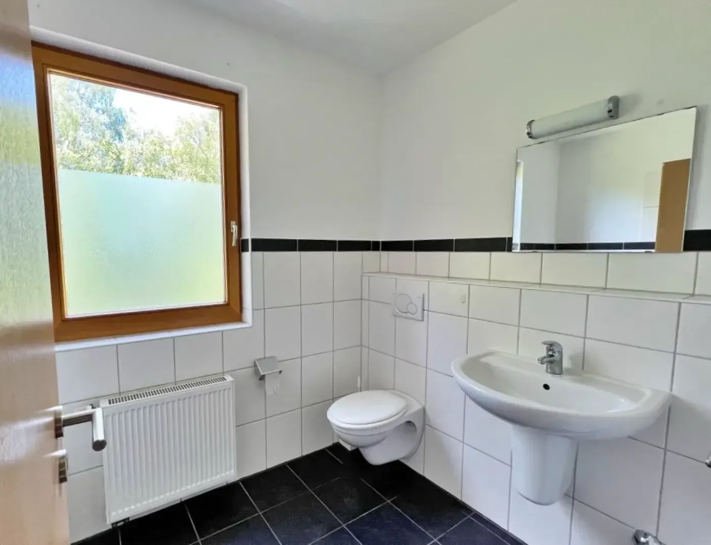 Ein weiß gefliestes Badezimmer mit Standwaschbecken, wandhängender Toilette, Heizkörper unter einem Milchglasfenster und einem Wandspiegel.