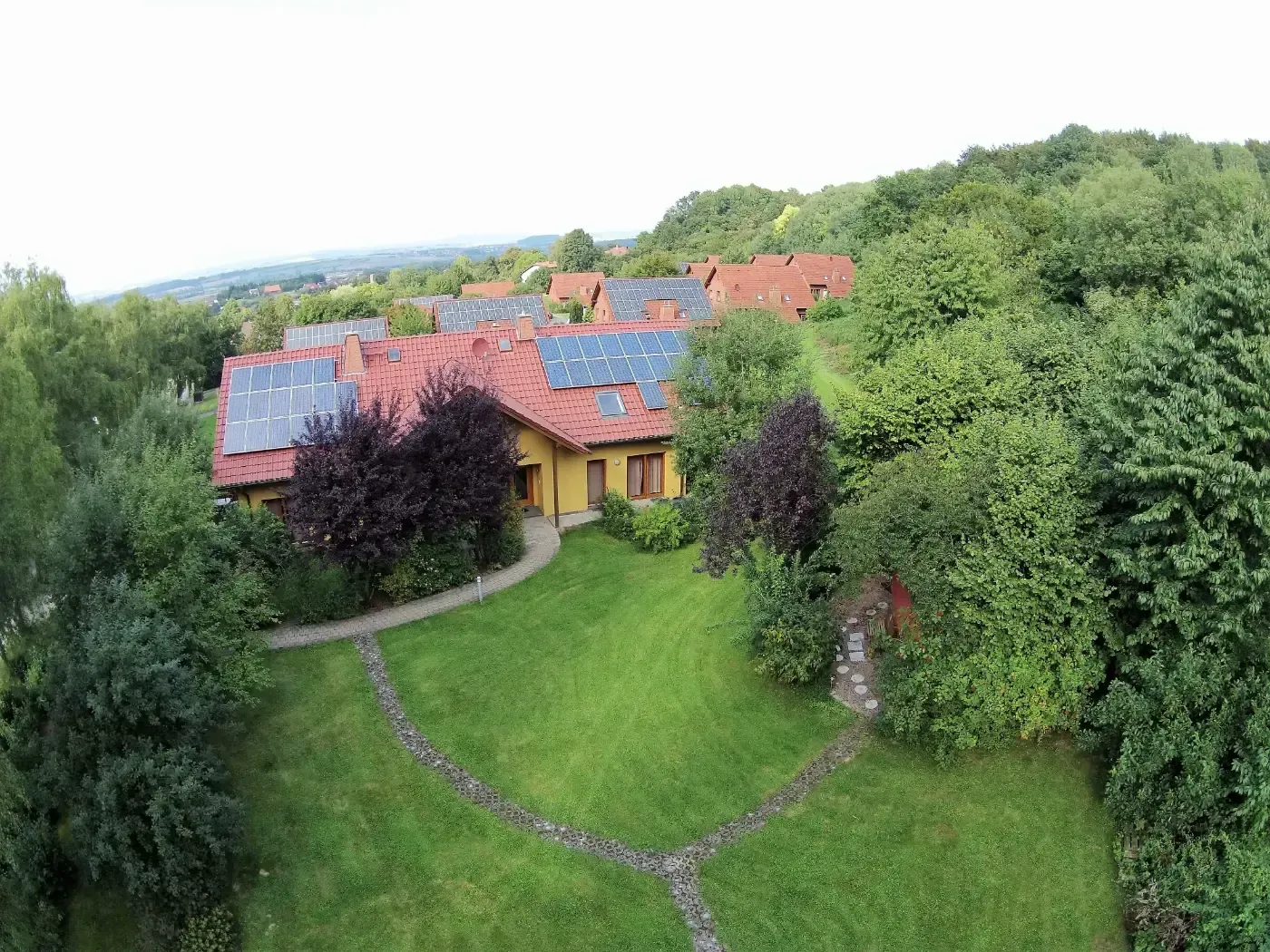 Luftaufnahme eines gelben Hauses mit rötlichem Dach und Solarpaneelen, umgeben von einem grünen Rasen und dichten Bäumen.