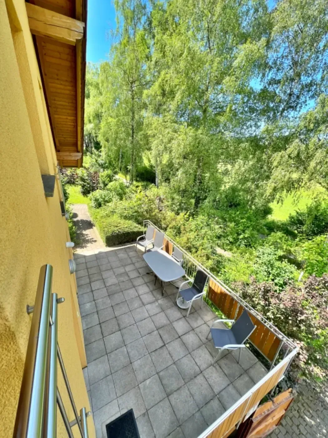 Blick von einem gelb getäfelten Balkon auf eine Terrasse mit Tisch, Stühlen und üppig grünen Bäumen im Hintergrund.
