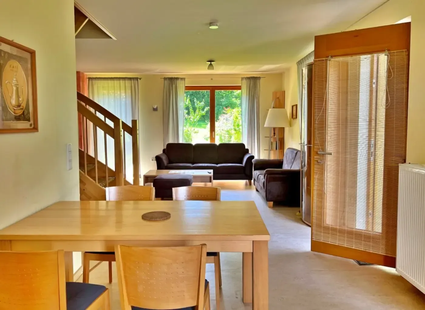 Im Vordergrund befindet sich ein Essbereich, der zu einem Wohnzimmer mit einem braunen Sofa, Stühlen und einer Treppe in einem hellen Haus führt.