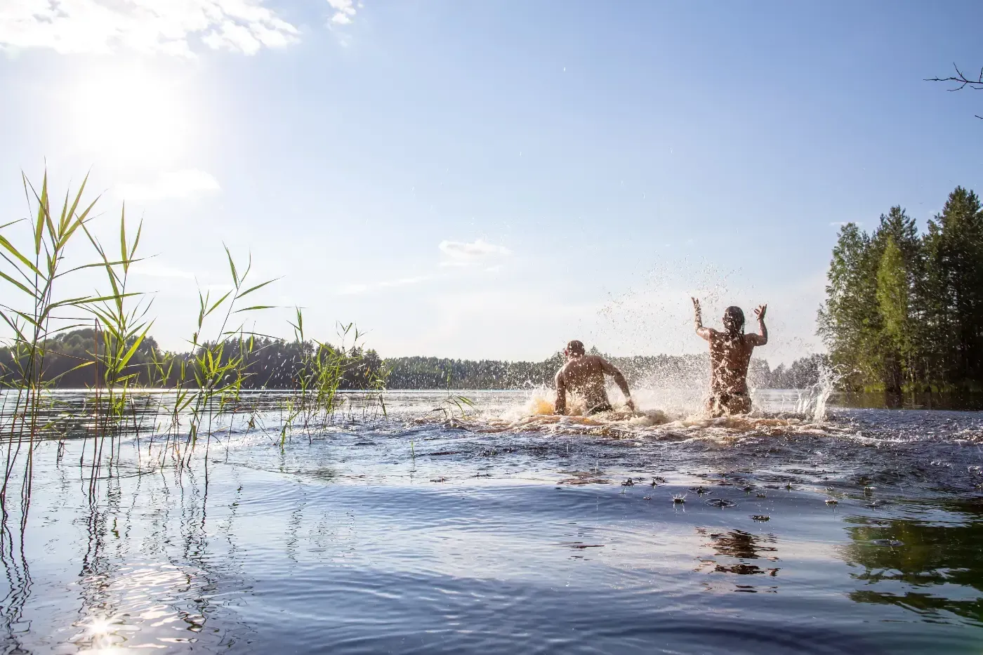 Zwei Personen springen in einen sonnenbeschienenen See und erzeugen dabei einen großen Platscher; im Hintergrund ist das Grün eines Waldes zu sehen.
