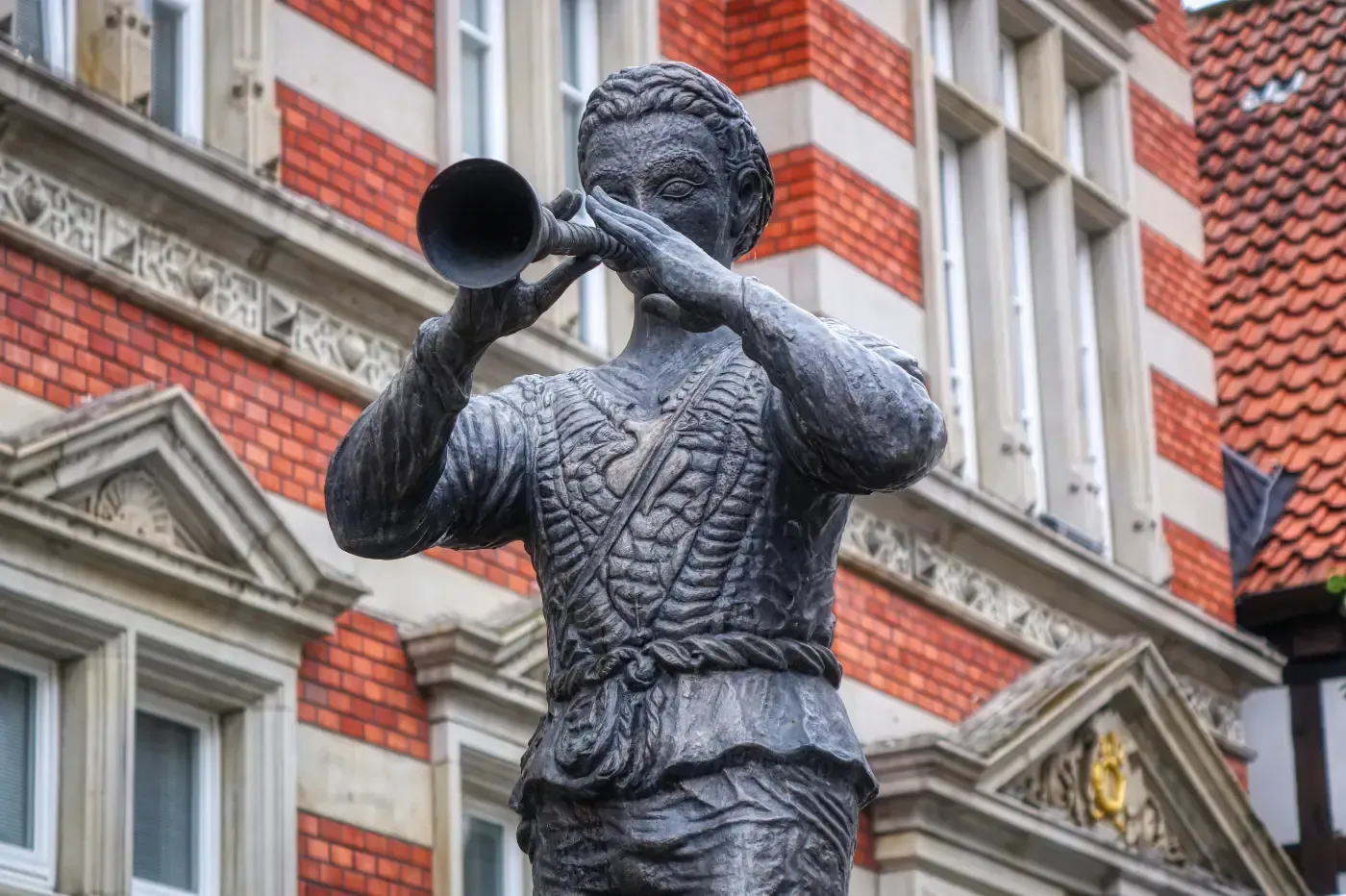 Eine Bronzestatue eines Trompeters vor einem roten Backsteingebäude mit architektonischen Details.