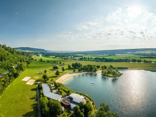 Luftaufnahme eines Sees mit Sandstrand, einem modernen Gebäudekomplex am Ufer und üppig grünen Feldern unter strahlendem Sonnenschein.