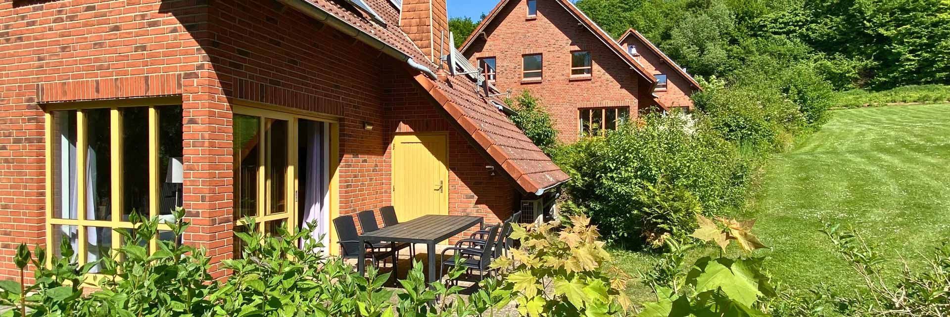 Rote Backsteinhäuser mit gelben Fensterrahmen stehen an einem grasbewachsenen Hang neben einer Terrasse mit Sitzgelegenheiten im Freien.