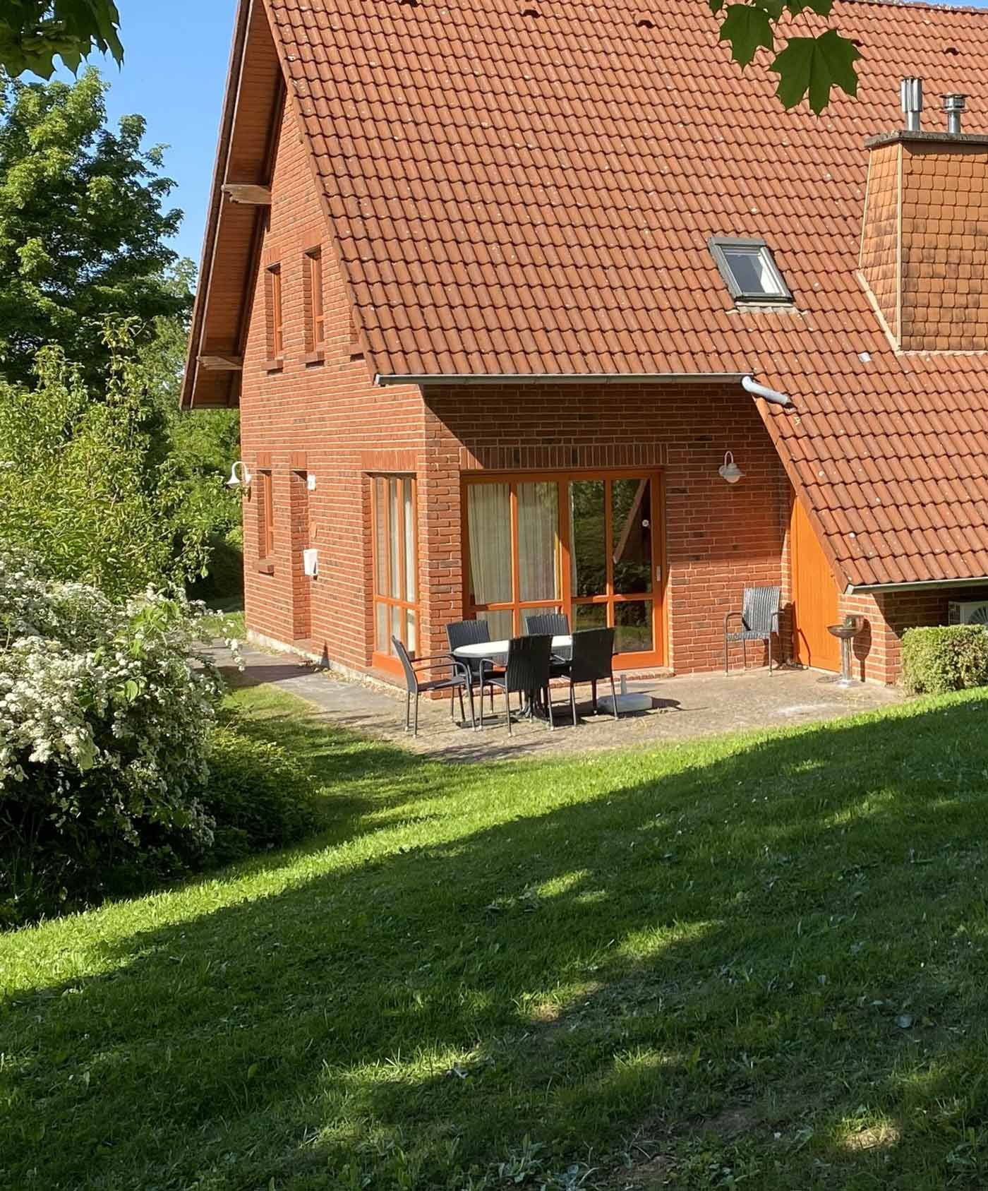 Ein Haus aus roten Ziegeln mit Ziegeldach, Gartenmöbel auf einer Betonterrasse und eine abfallende Rasenfläche im Vordergrund.