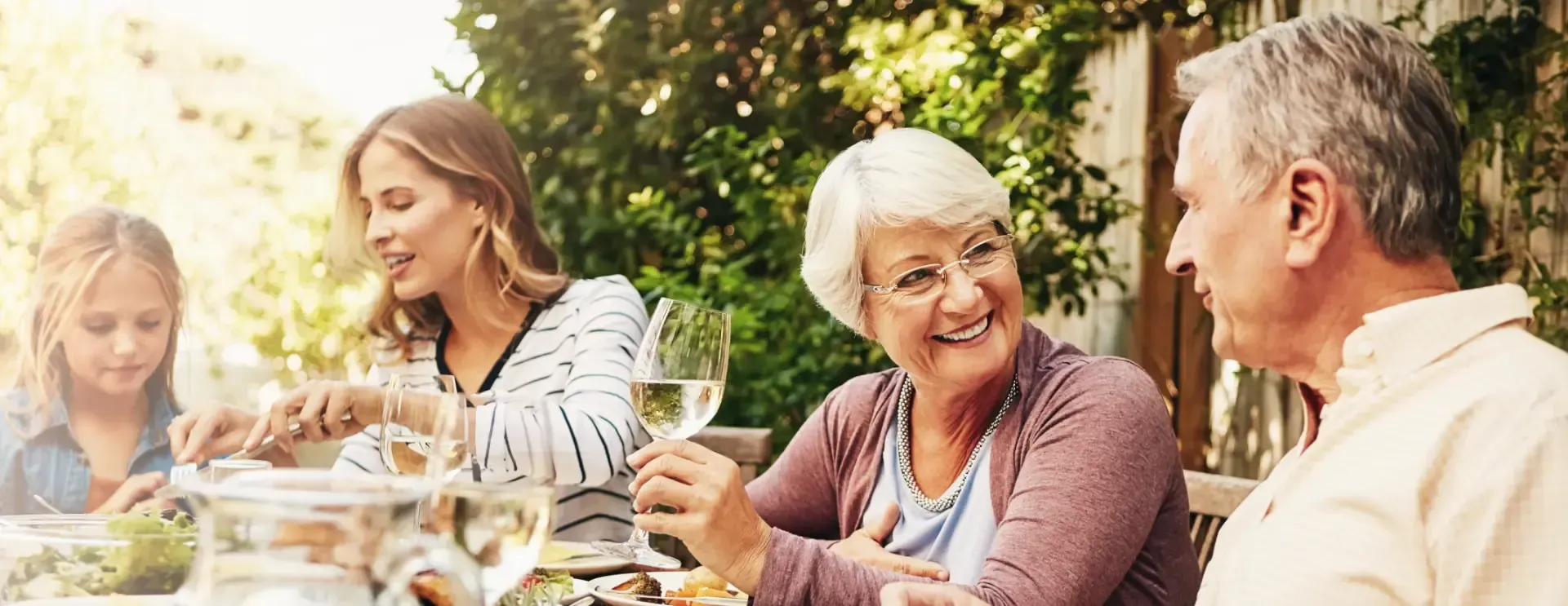 Eine Familie genießt eine Mahlzeit im Freien an einem sonnigen Terrassentisch und unterhält sich lachend beim Essen.