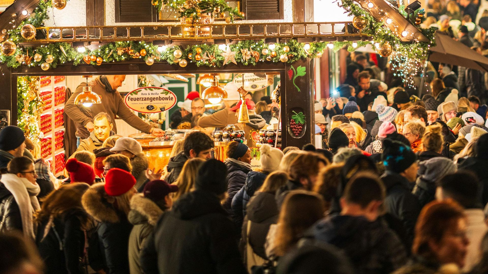 Eine Menschenmenge versammelt sich an einem warm beleuchteten, mit festlichem Grün und Ornamenten geschmückten Weihnachtsmarktstand im Freien.