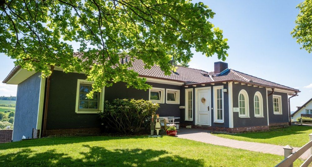Eingeschossiges Haus mit dunkler Fassade, weißen Zierelementen und einem grünen Rasen unter belaubten Bäumen im Sonnenlicht