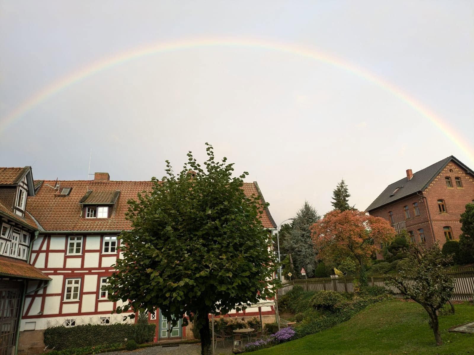 Dorfhäuser und Bäume unter einem schwachen Regenbogen an einem bedeckten Himmel