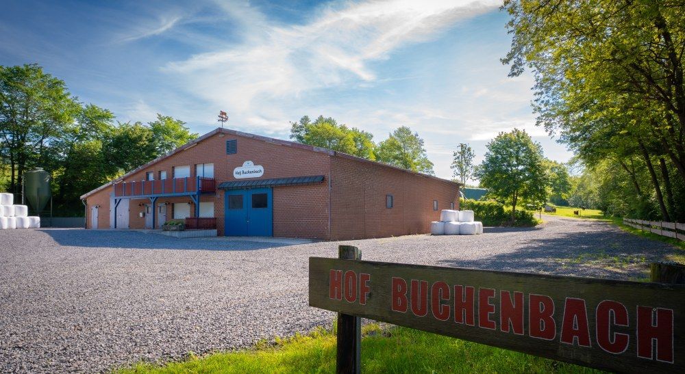 Backsteingebäude mit blauen Türen und Kiesplatz, im Vordergrund ein Schild mit der Aufschrift „Hof Buchenbach“