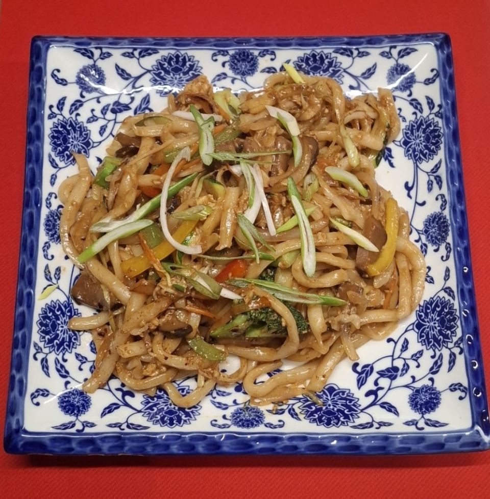 Udon-Nudeln mit Gemüse und geschnittenen Frühlingszwiebeln auf einem blau-weiss gemusterten Teller.