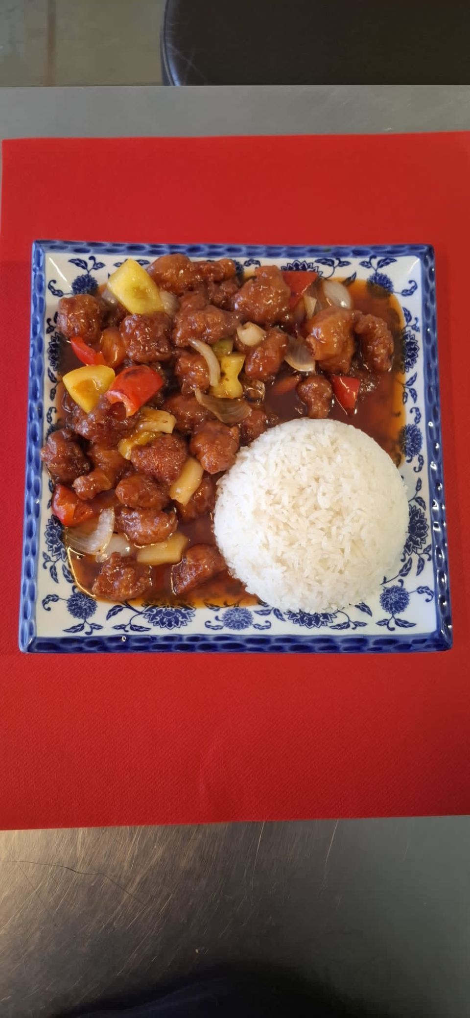 Süss-saures Schweinefleisch mit Reis auf einem quadratischen Teller mit blau-weissem Muster, auf einem roten Platzdeckchen.