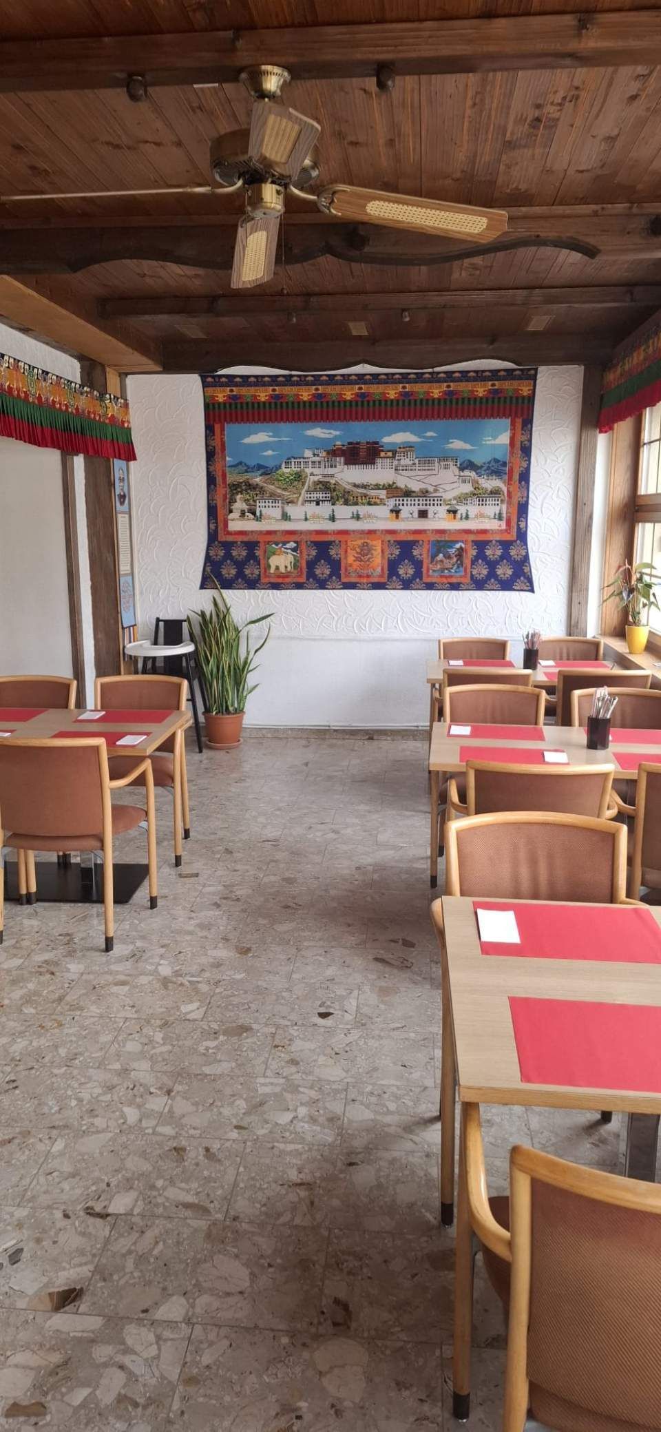 Restaurantinterieur mit Tischen, roten Platzsets und einem Wandteppich. Deckenventilator und Steinboden.