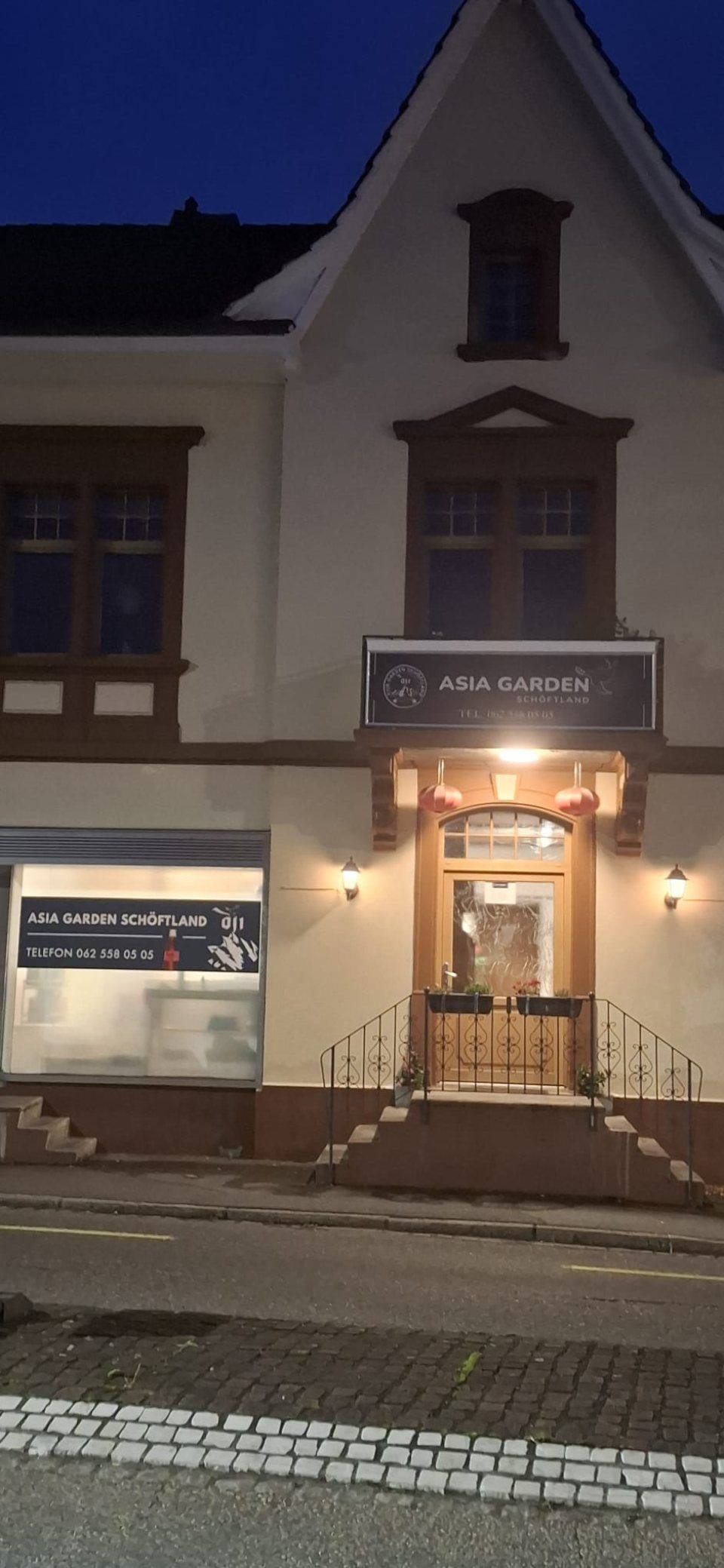 Aussenansicht eines Gebäudes bei Nacht. Eine beleuchtete Tür, ein Fenster und ein Schild sind zu sehen.