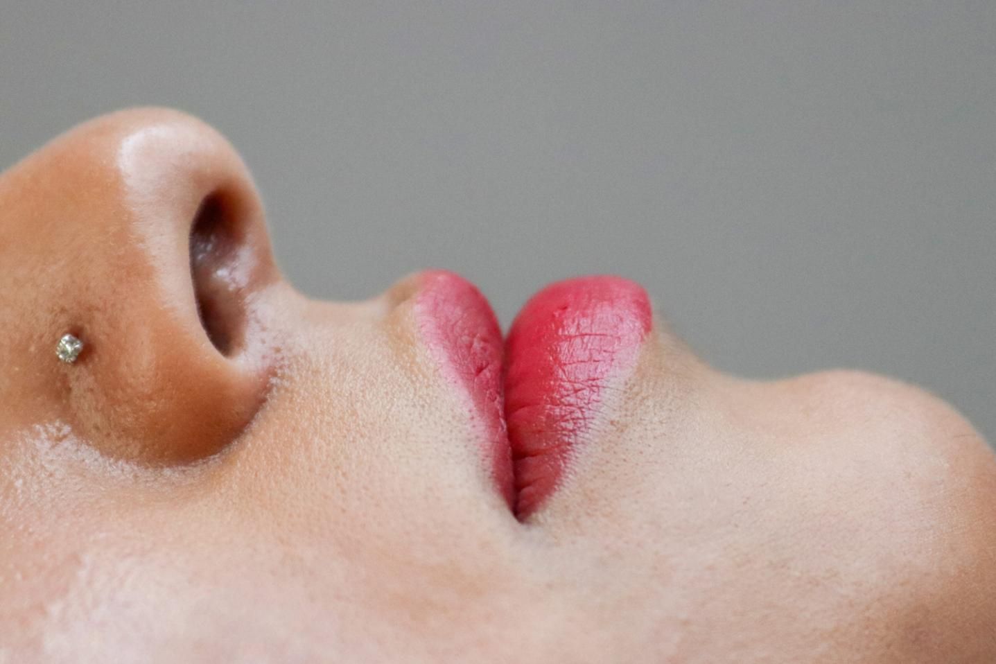 Primer plano de un rostro de perfil con un piercing en la nariz y labios rosados sobre un fondo gris.