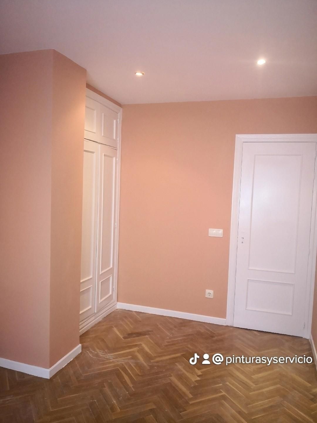 Habitación vacía de color melocotón con puertas blancas, suelo de madera en espiga y luces en el techo.