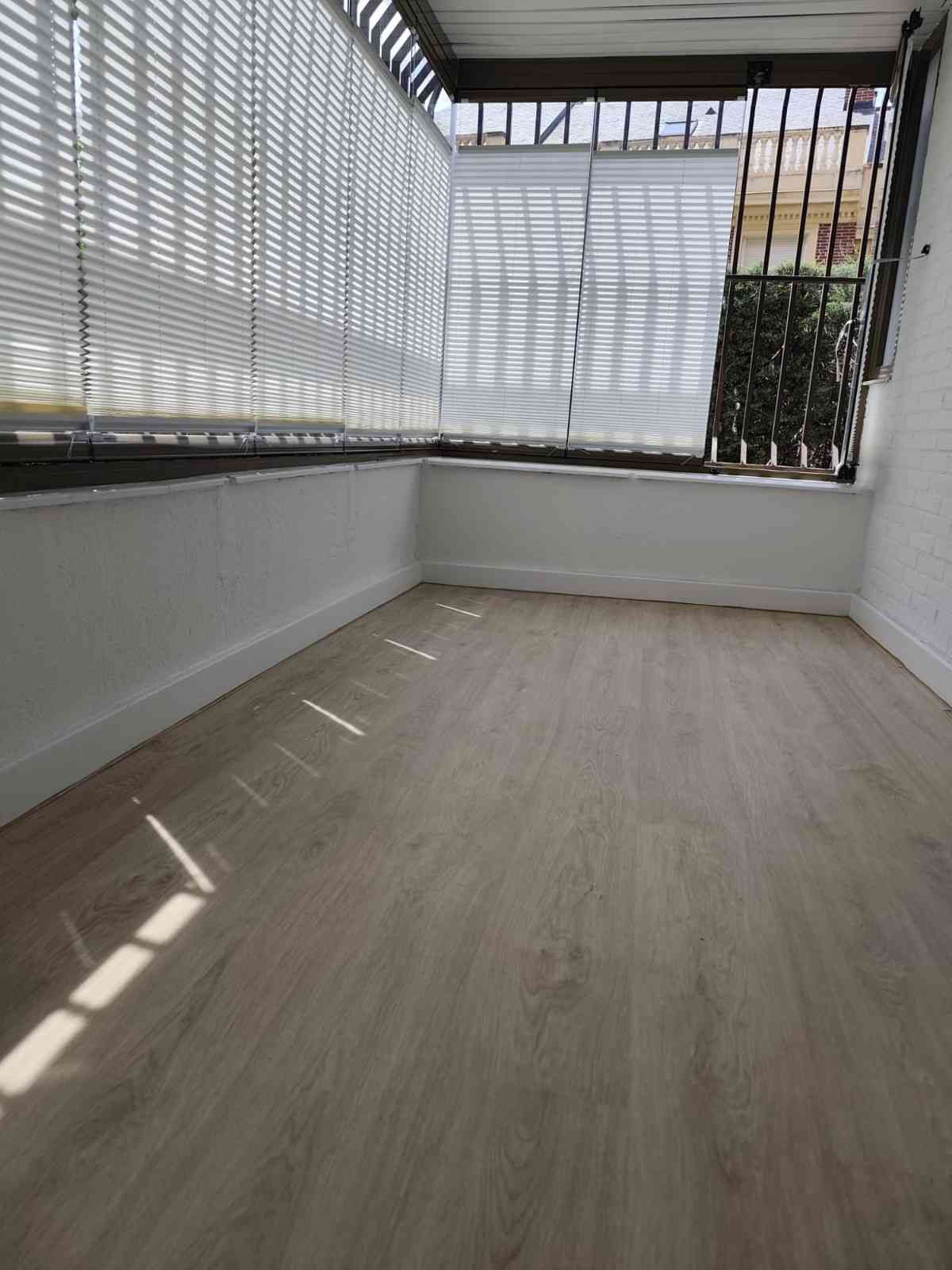 Un luminoso y vacío solárium con persianas blancas, suelo de madera clara y luz solar que entra a raudales por las ventanas.