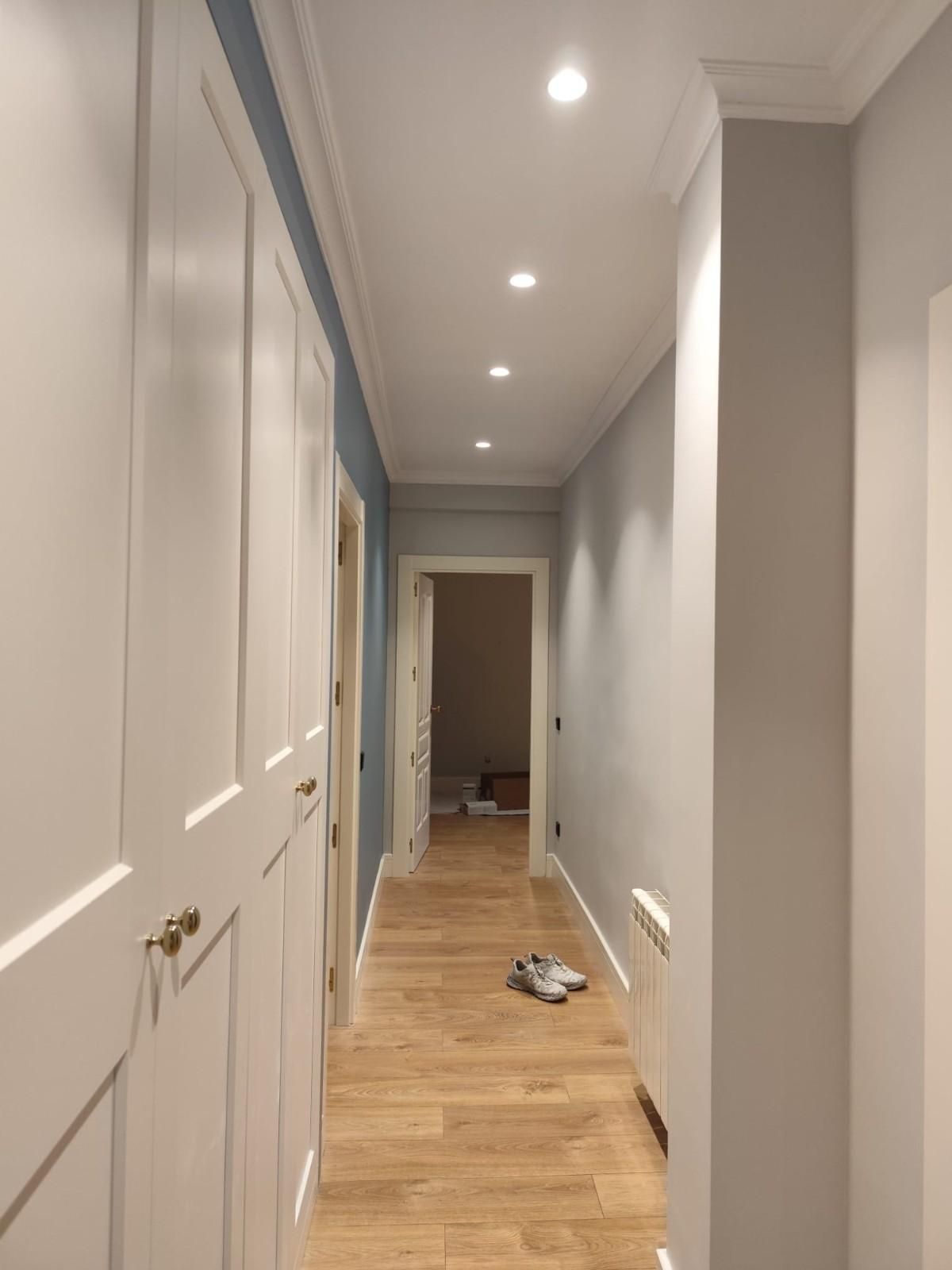 Pasillo estrecho con paredes blancas, suelo de madera, luces empotradas y puertas a ambos lados.