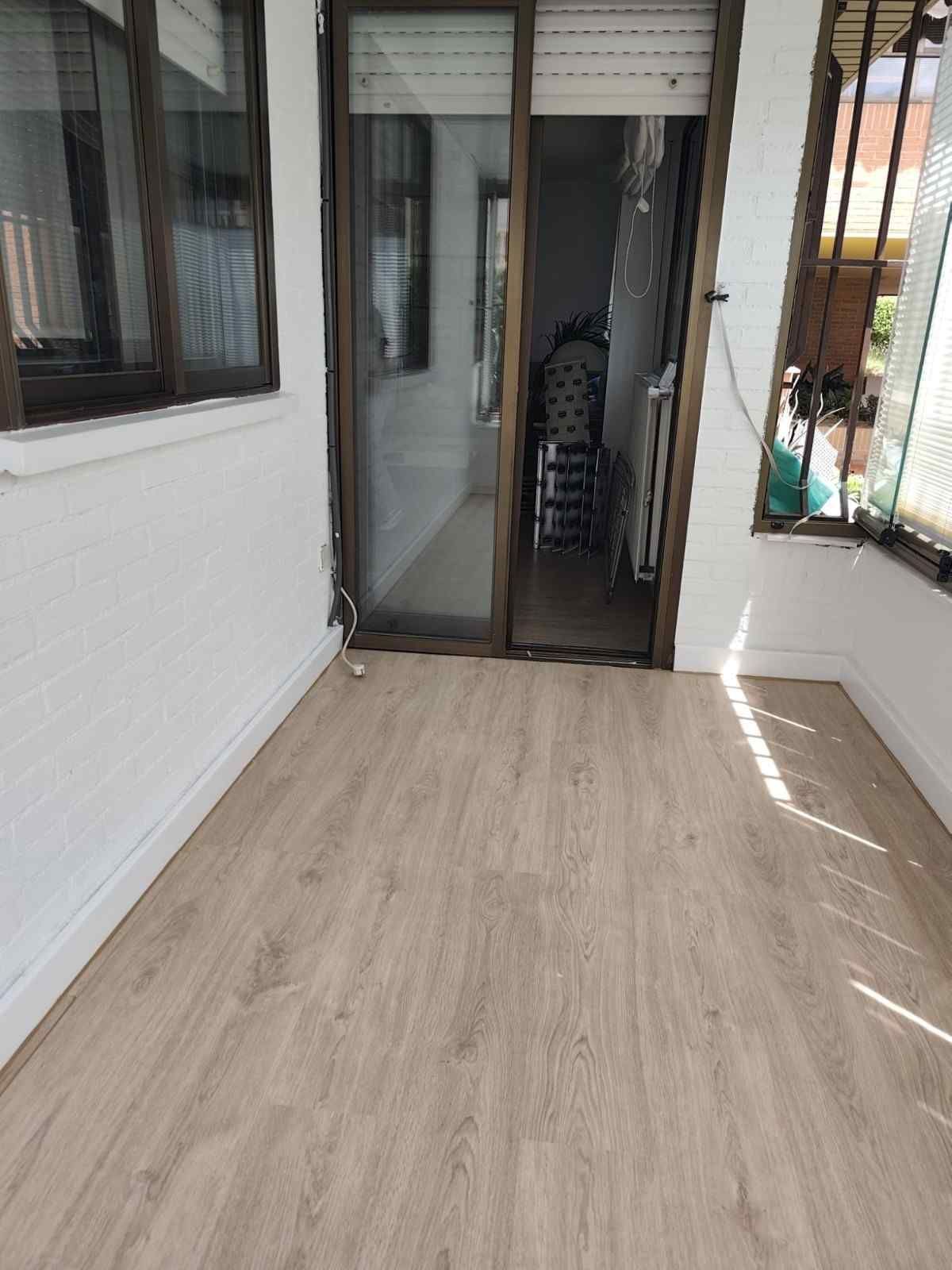 Balcón estrecho y cerrado con suelo de madera clara, paredes blancas y una puerta corredera de cristal en el extremo opuesto.