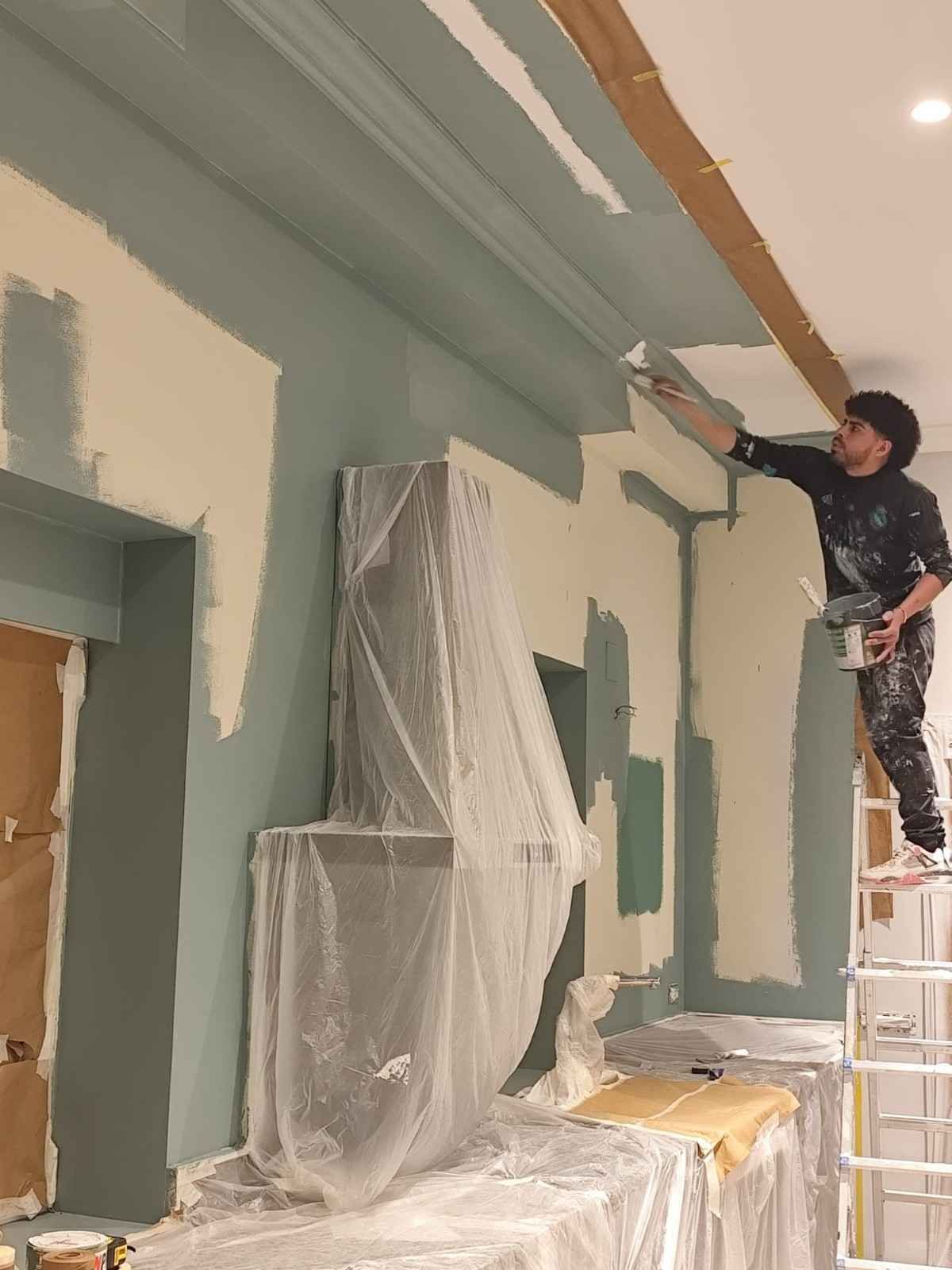 Pintor subido a una escalera pintando el borde del techo en una habitación con muebles tapizados y paredes verdes.