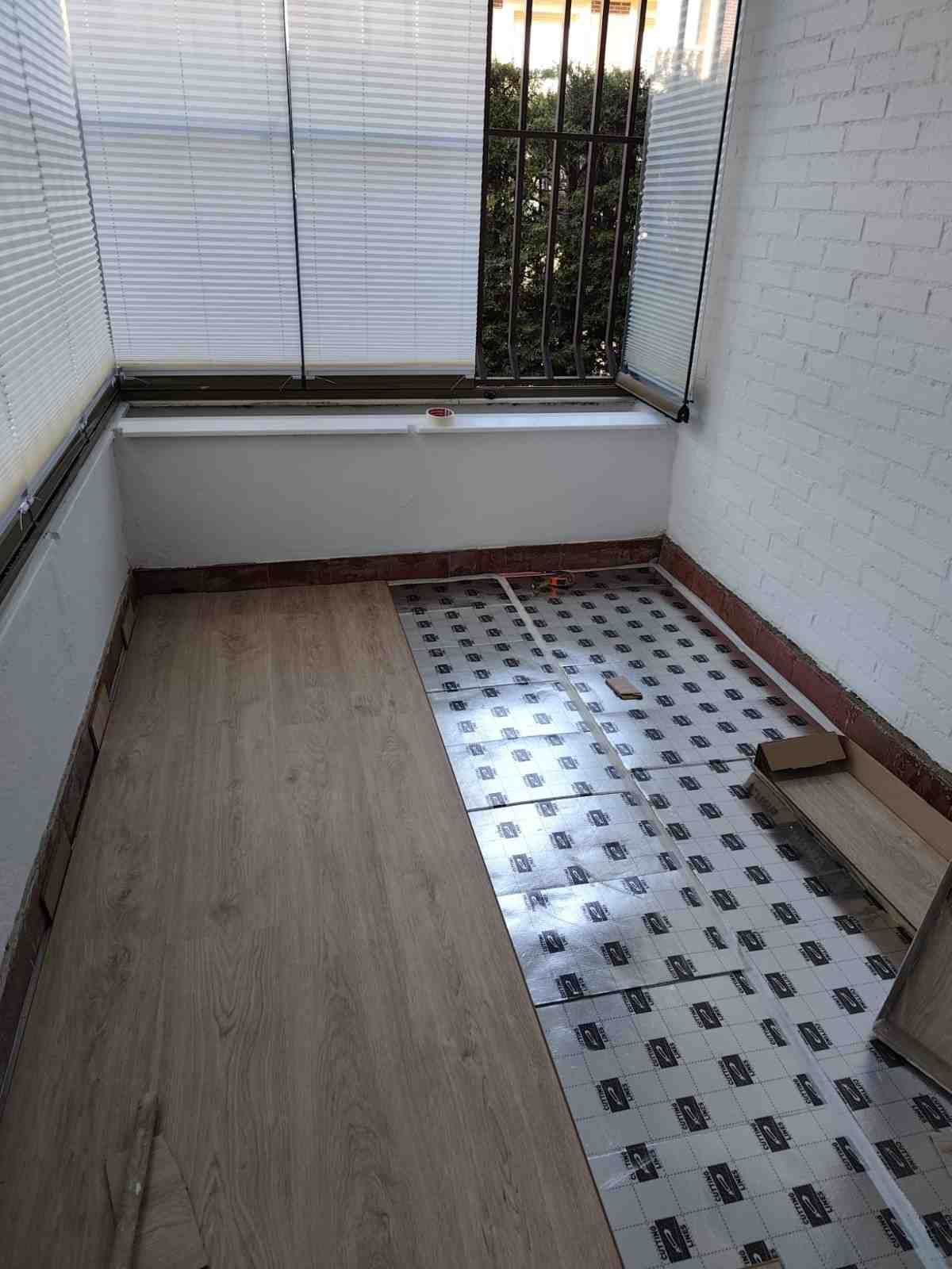 Pequeño balcón con suelo de madera, franja de azulejos, paredes blancas y barandilla metálica.