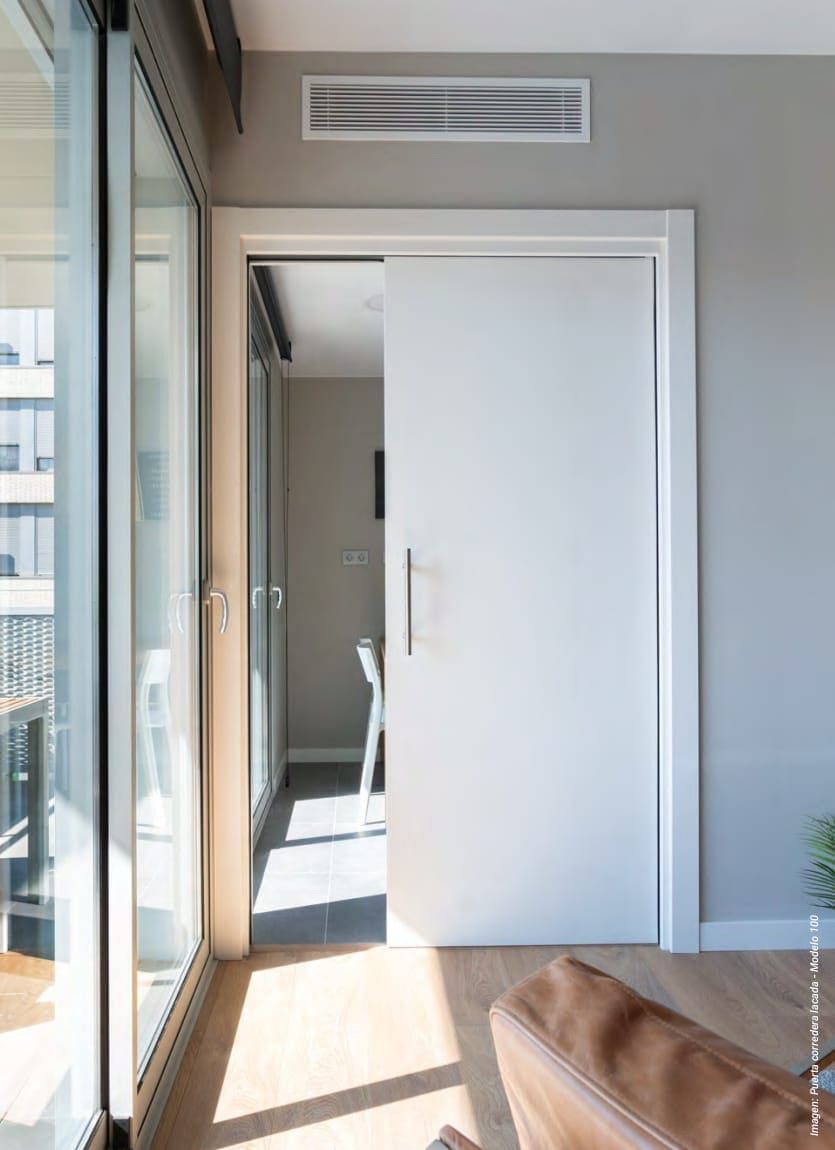 Una puerta corredera blanca integrada en una pared de tonos neutros, con una rejilla de ventilación lineal en el techo y una vista parcial de una habitación luminosa al otro lado.