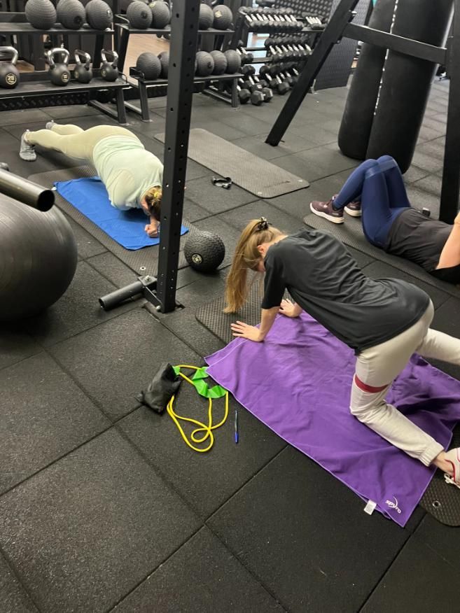 Hombre haciendo remos con mancuernas en un banco, guiado por un entrenador en un gimnasio.