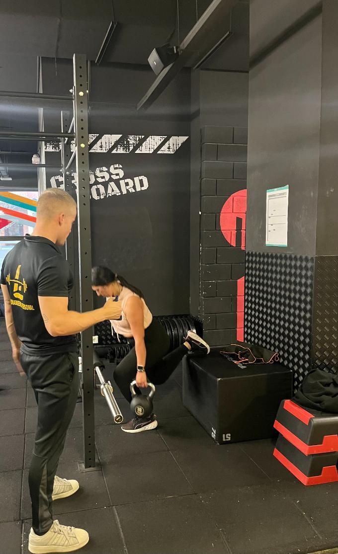 Mujer haciendo remos con mancuernas en un banco, guiada por un entrenador en un gimnasio.