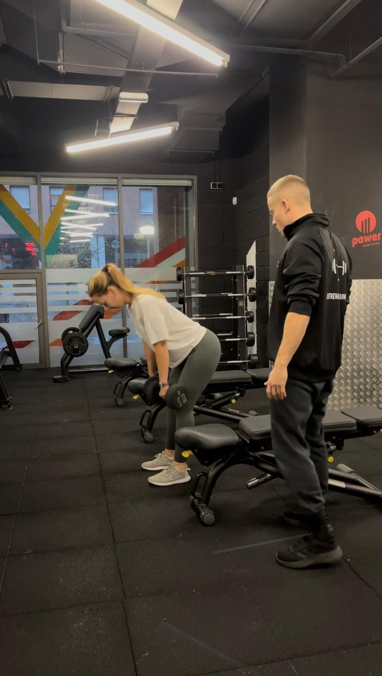 Mujer haciendo remos con mancuernas en un banco, guiada por un entrenador en un gimnasio.