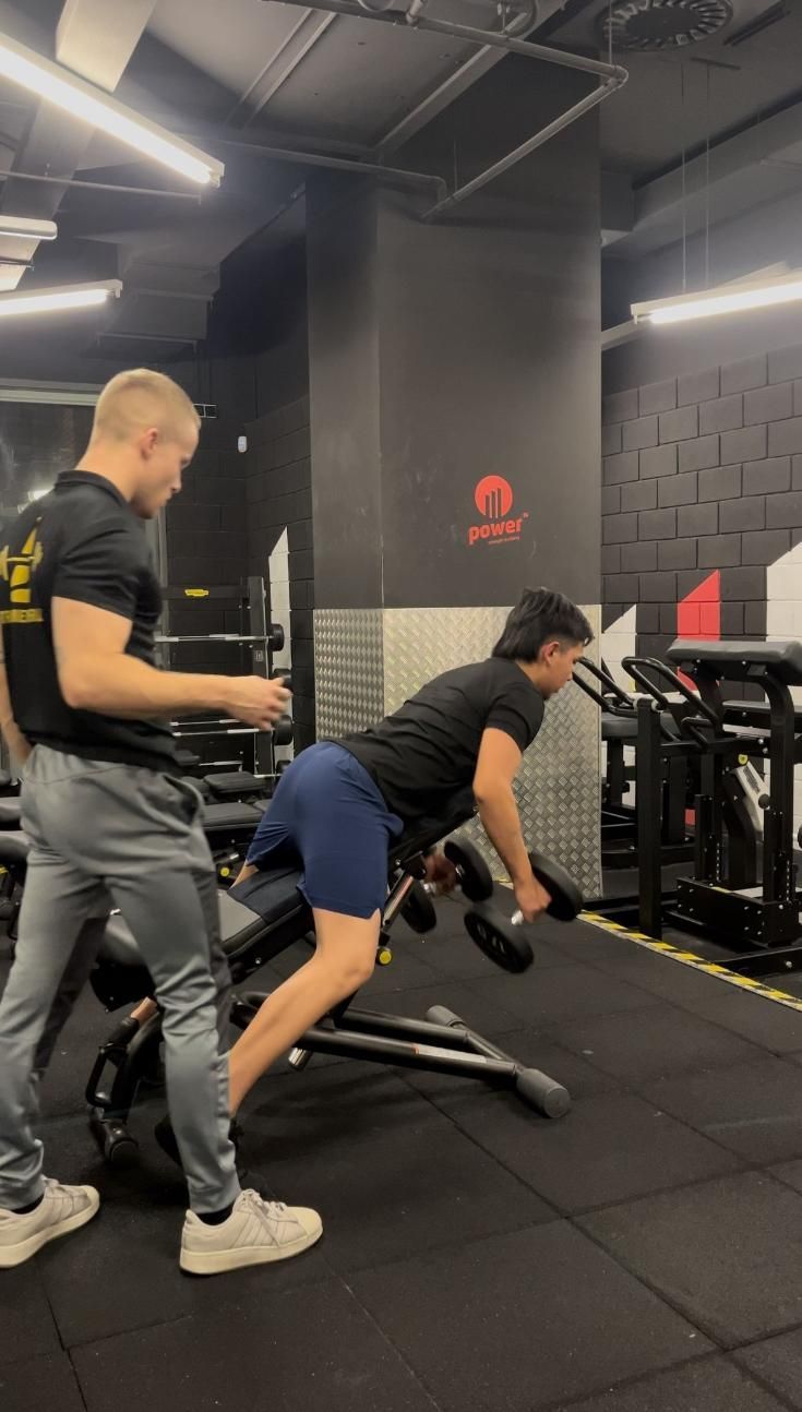 Hombre haciendo remos con mancuernas en un banco, guiado por un entrenador en un gimnasio.
