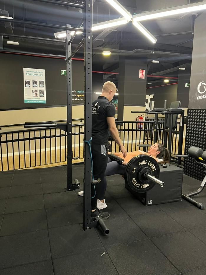 Un entrenador observa a una persona haciendo press de banca en un gimnasio. Barra negra con pesas.