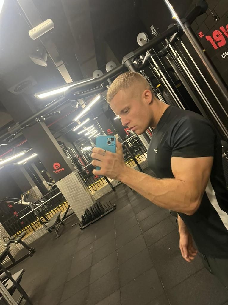 Hombre en el gimnasio tomándose una selfi frente al espejo. Lleva camiseta negra y se le ve el brazo musculoso. 