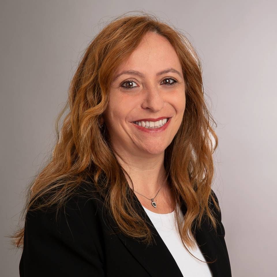 Eine Frau mit langen, rotbraunen Haaren lächelt. Sie trägt einen schwarzen Blazer über einem weißen Hemd. Der Hintergrund ist grau.