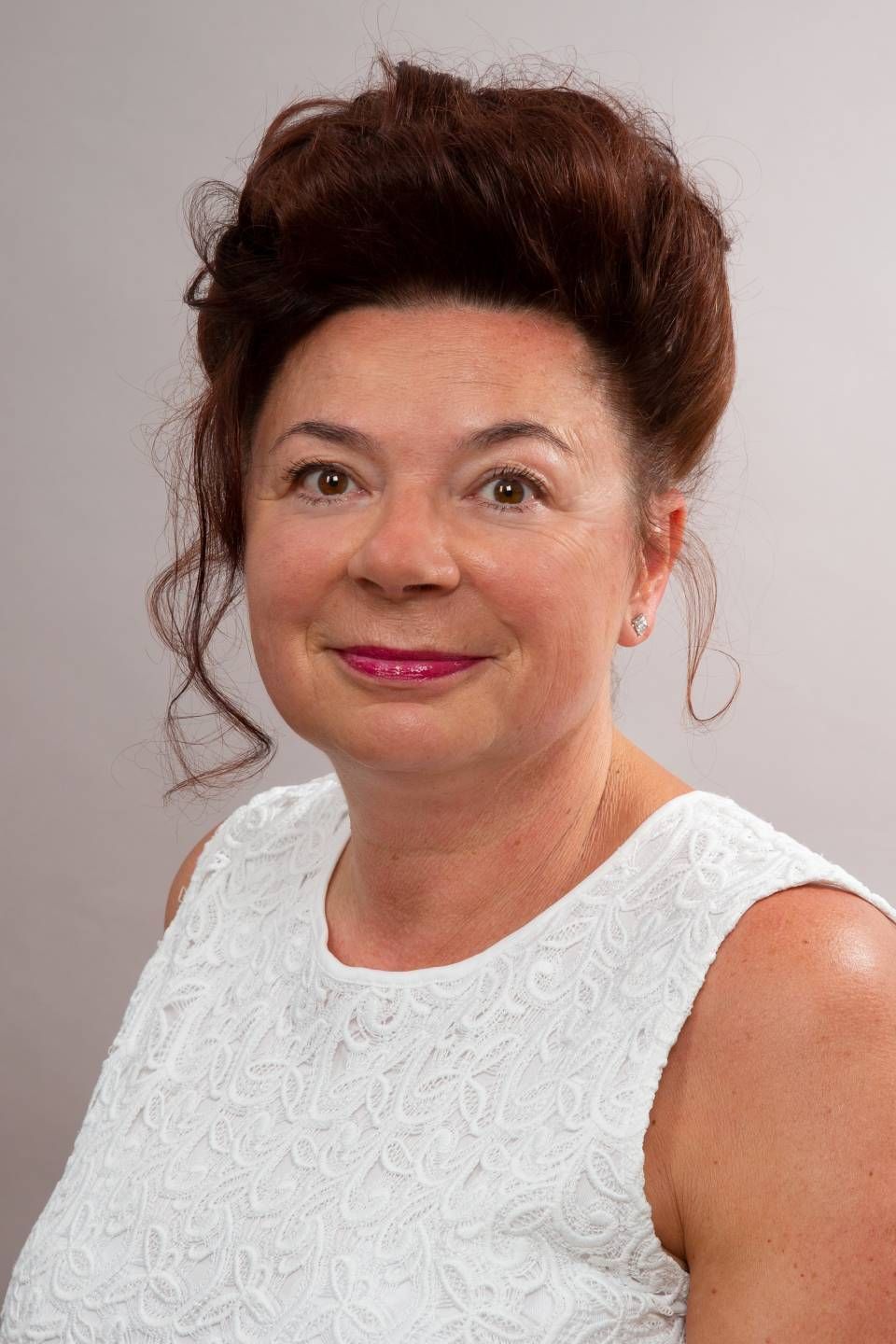 Eine Frau mit dunklen, lockigen Haaren und einem weißen, ärmellosen Top lächelt vor einem neutralen Hintergrund in die Kamera.