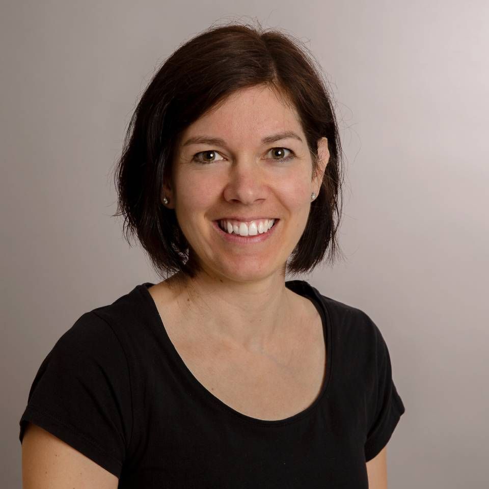 Eine Frau mit kurzen braunen Haaren lächelt in einem schwarzen T-Shirt vor einem hellgrauen Hintergrund.