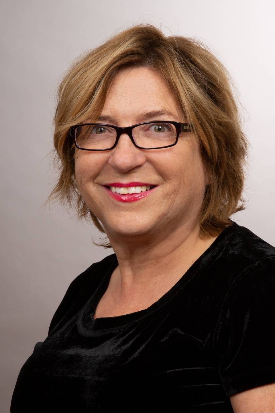 Frau mit Brille, lächelnd und mit rotem Lippenstift, in einem schwarzen Oberteil, vor grauem Hintergrund.