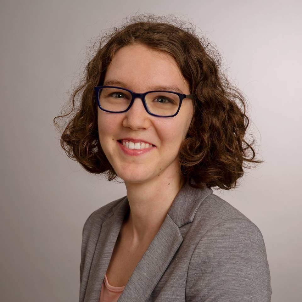 Frau mit lockigem braunem Haar, Brille und grauem Blazer lächelt.