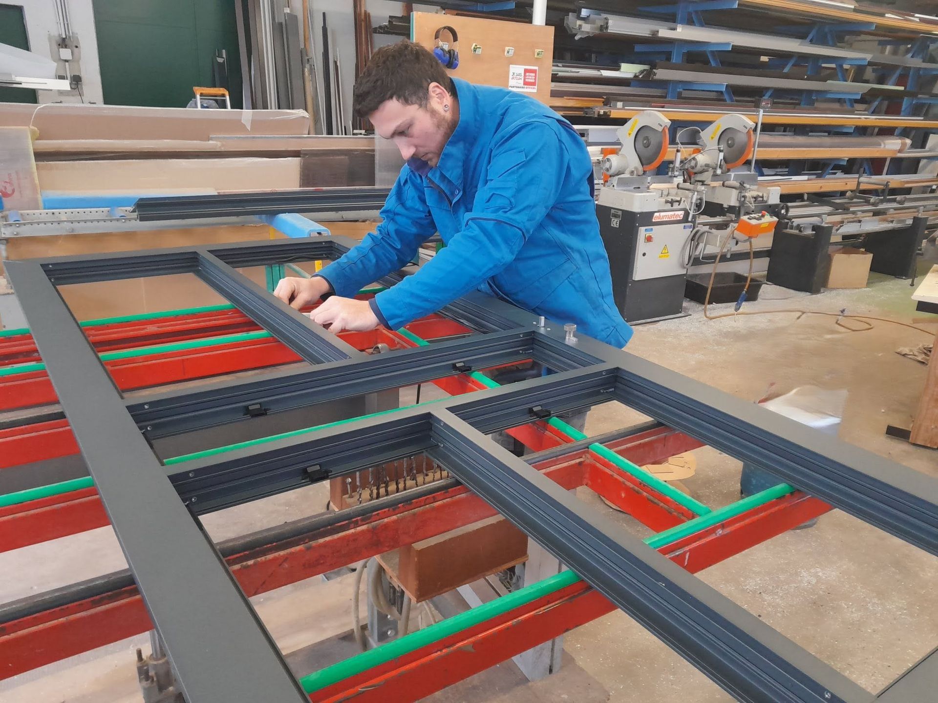 Un ouvrier en veste bleue assemble une structure métallique dans un atelier où se trouvent des outils et des machines.