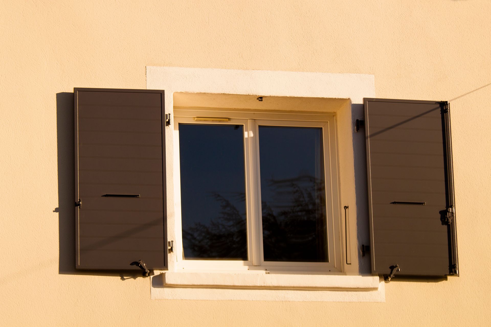 Fenêtre à cadre blanc et volets foncés sur un mur extérieur beige.