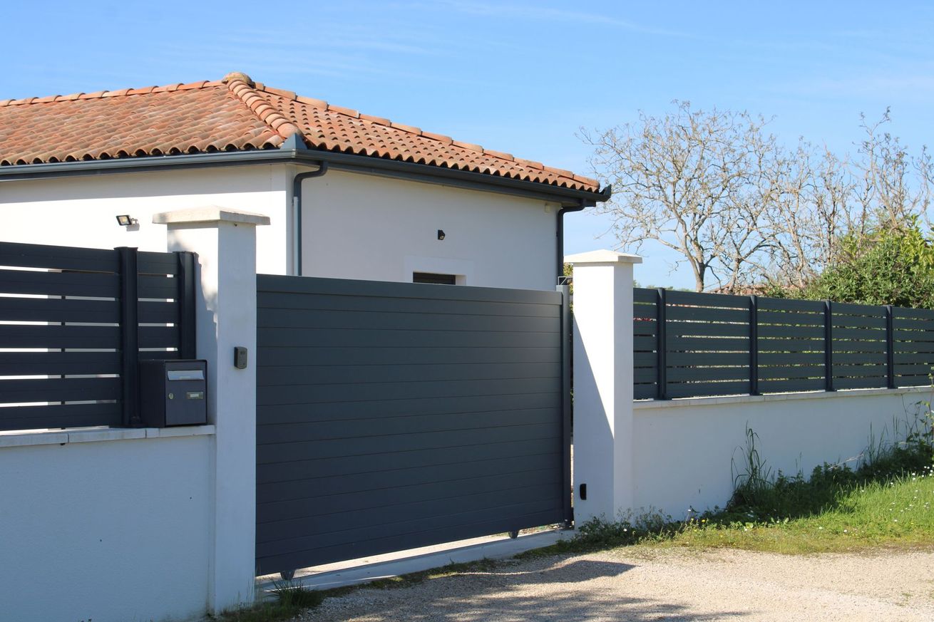 Maison moderne aux murs blancs, portail et clôture gris foncé.