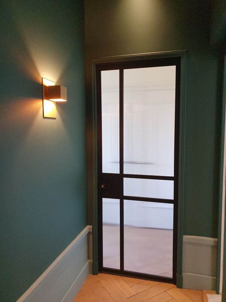 Un couloir aux murs turquoise, une porte vitrée à cadre noir et un luminaire mural.