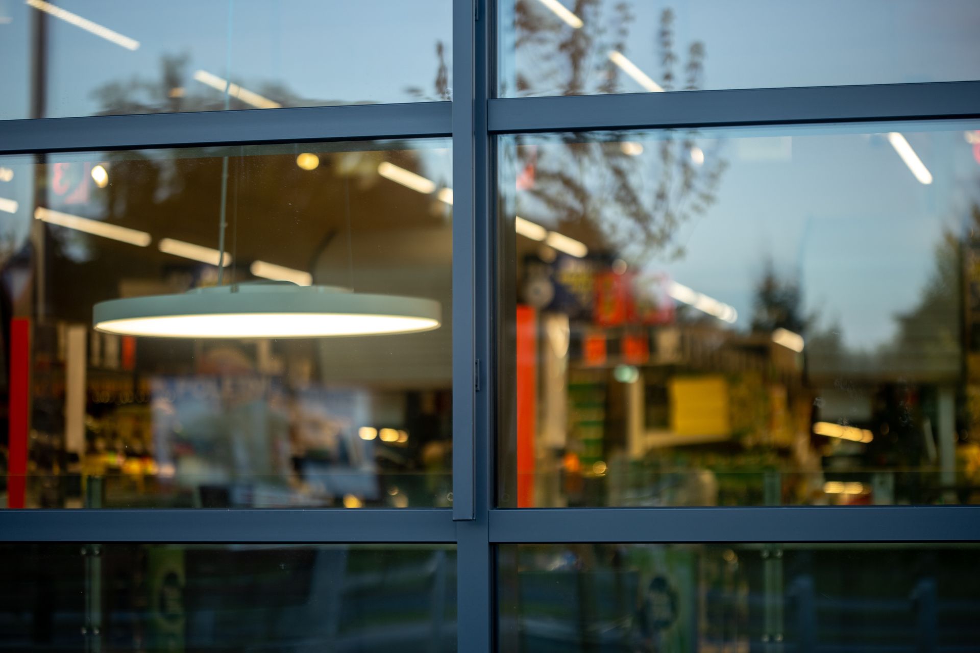Reflets dans une vitrine : intérieur avec un luminaire circulaire allumé et scène extérieure floue de verdure et d’étalages de magasins.