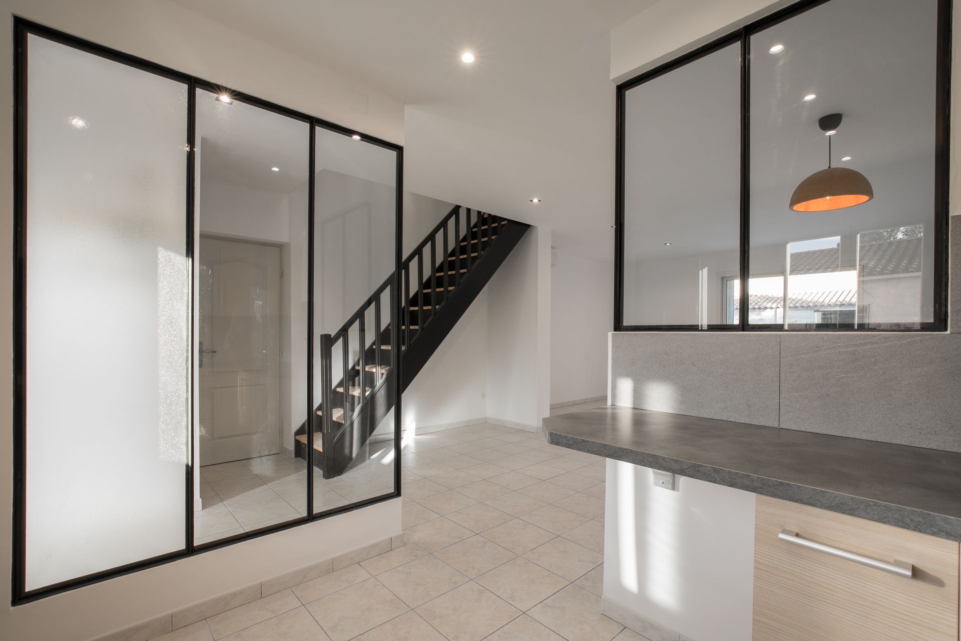 Un intérieur moderne avec une cloison vitrée à cadre noir, un escalier noir et un comptoir de cuisine avec des armoires en bois clair.