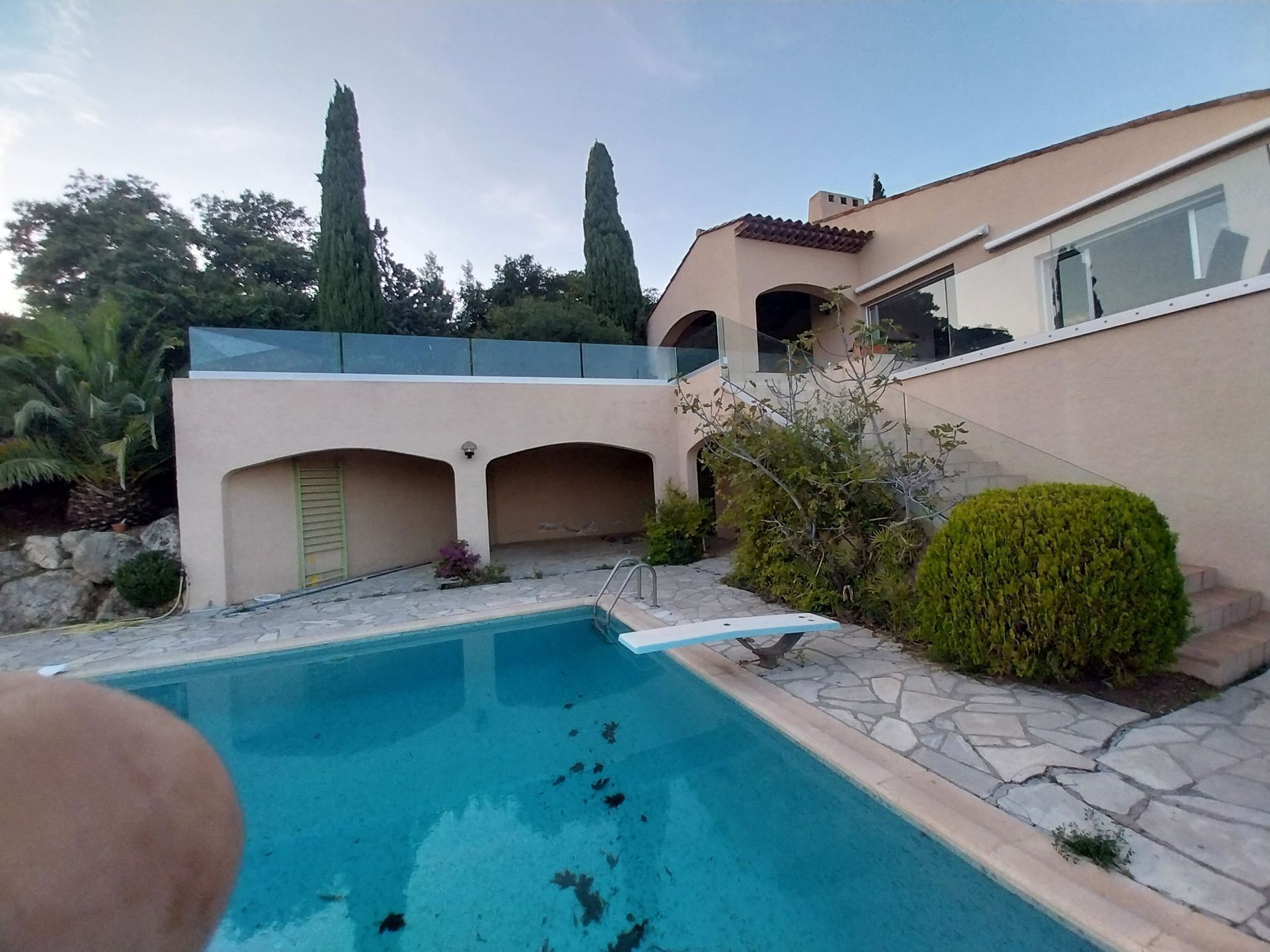 Piscine avec plongeoir devant une maison claire. Arbres et ciel bleu en arrière-plan.